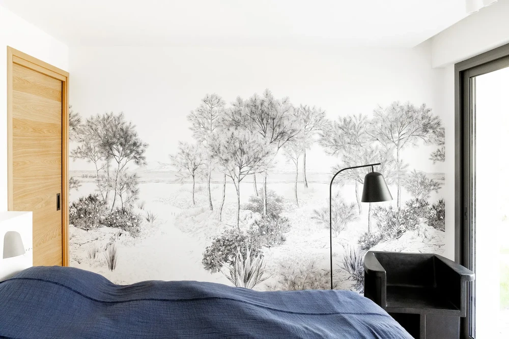 Chambre contemporaine avec papier peint panoramique représentant un paysage pastoral en noir et blanc, lit aux draps bleus, fauteuil design noir et lampadaire arqué